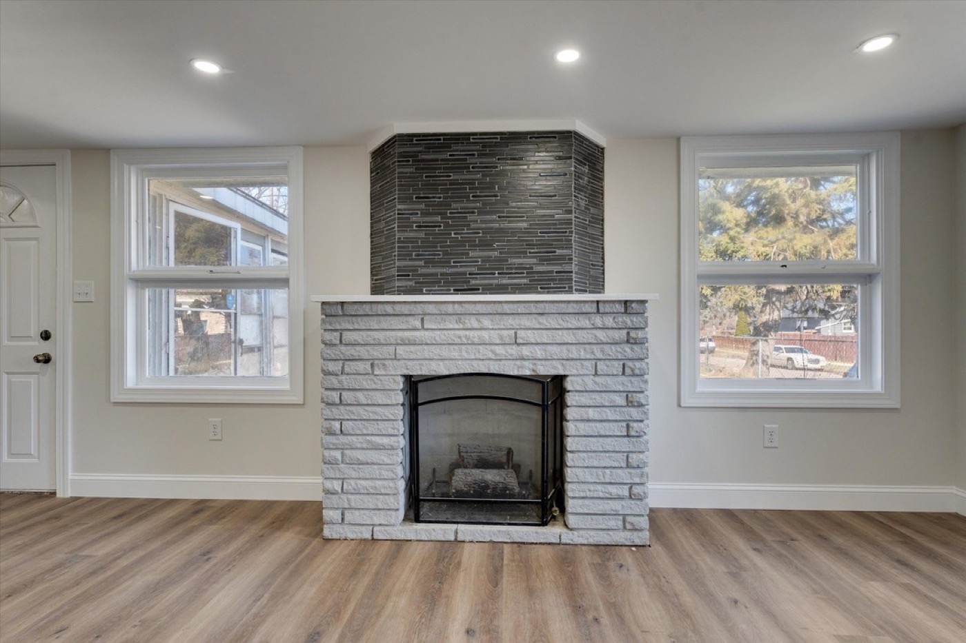 Living room after renovation: whitewashed brick fireplace, dark tiled chimney breast, wood floors, windows either side.