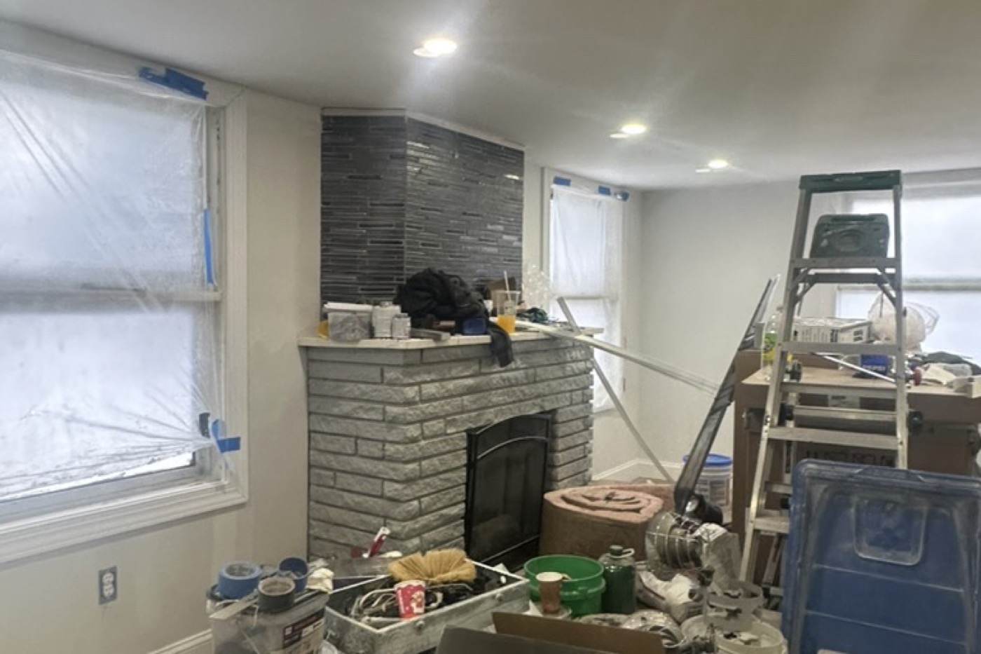 Living room during renovation: bare brick fireplace, masked windows, contractor materials on the floor.