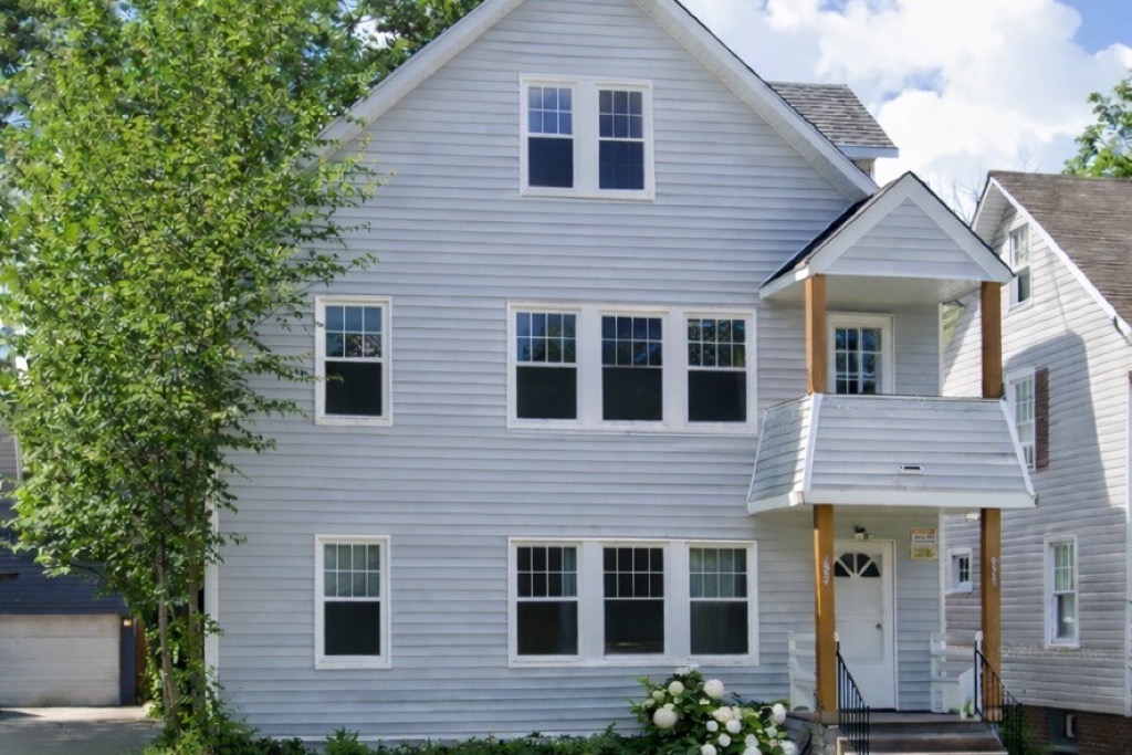 3220 Clarendon Road after renovation: grey lap-sided triplex with white trim, covered entry on wood posts, fresh landscaping and hydrangeas by the steps.