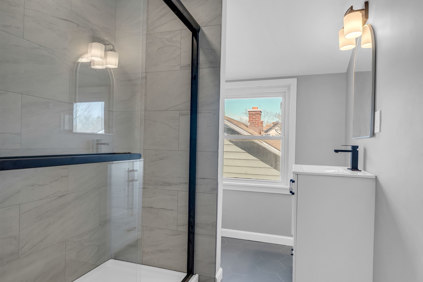 Upstairs bathroom after renovation: tiled glass shower on the left, window ahead, white vanity with black fixtures on the right, hexagon tile floor.