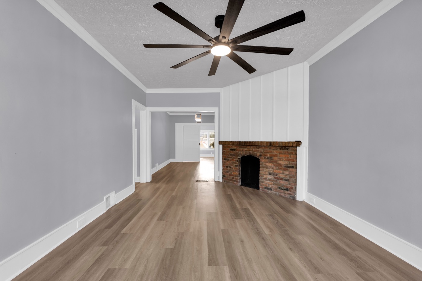 Living room after renovation: brick fireplace with white panelled chimney breast on the right, new plank flooring, ceiling fan, opening through to the dining room.