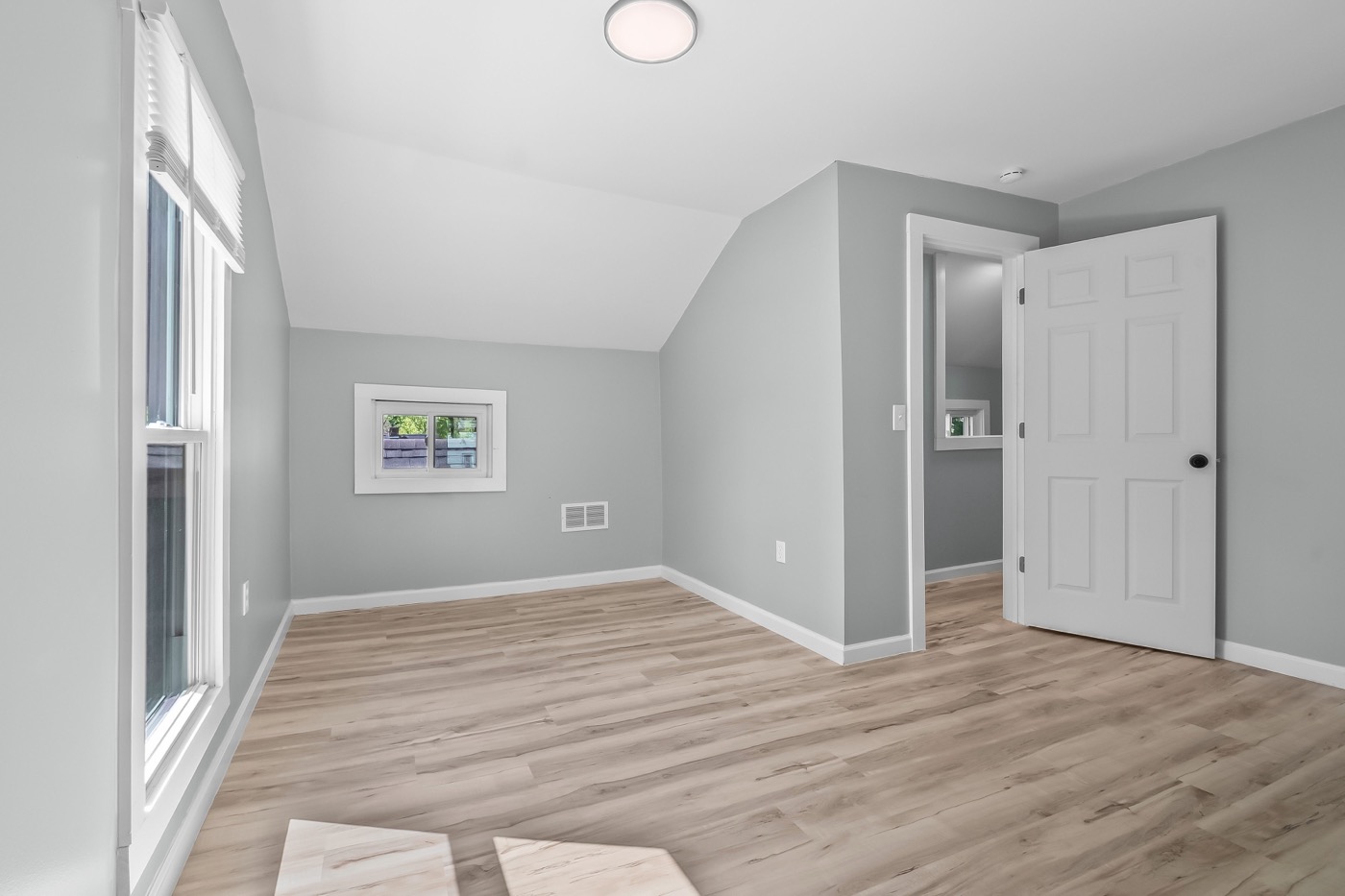 Bedroom after renovation: new drywall, grey paint, new plank flooring, ceiling light, and a window over the yard.