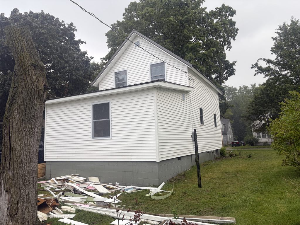 Side of the house with new white siding, new windows, standing-seam metal roof, and painted block foundation; old siding stacked on the lawn.