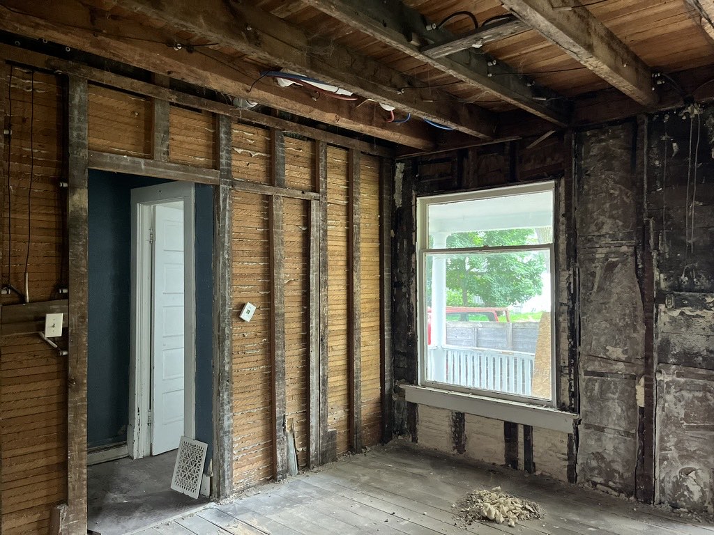 Room stripped to the original wood lath and framing, with a window looking onto the covered porch.
